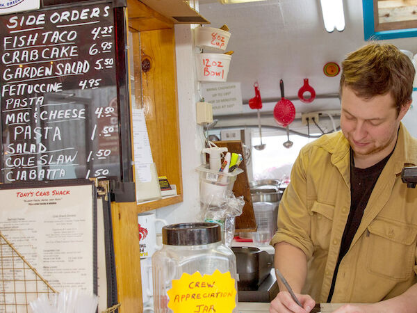 A person stands at a counter taking orders in a food place. A tip jar, menu board, and "Order Here" sign are visible.