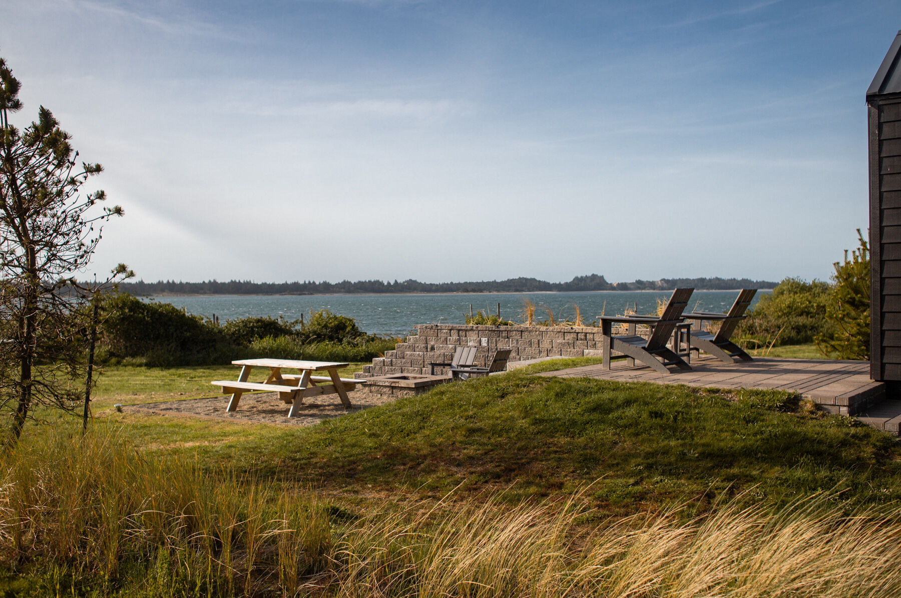A scenic view with a picnic table, chairs on a deck, and grassy area overlooking a body of water under a clear blue sky.