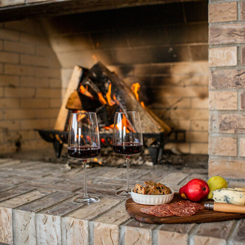 Two glasses of red wine by a fireplace, with a platter of cheese, salami, a pear, a pomegranate, and crackers on a wooden board.