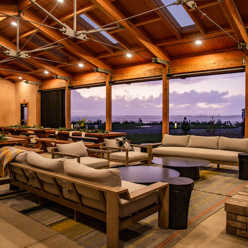 A spacious indoor area with wooden beams, sofas, and tables, overlooking a scenic sunset. Cozy and inviting setup.