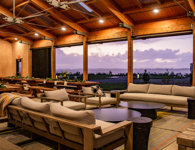 A spacious indoor area with wooden beams, sofas, and tables, overlooking a scenic sunset. Cozy and inviting setup.