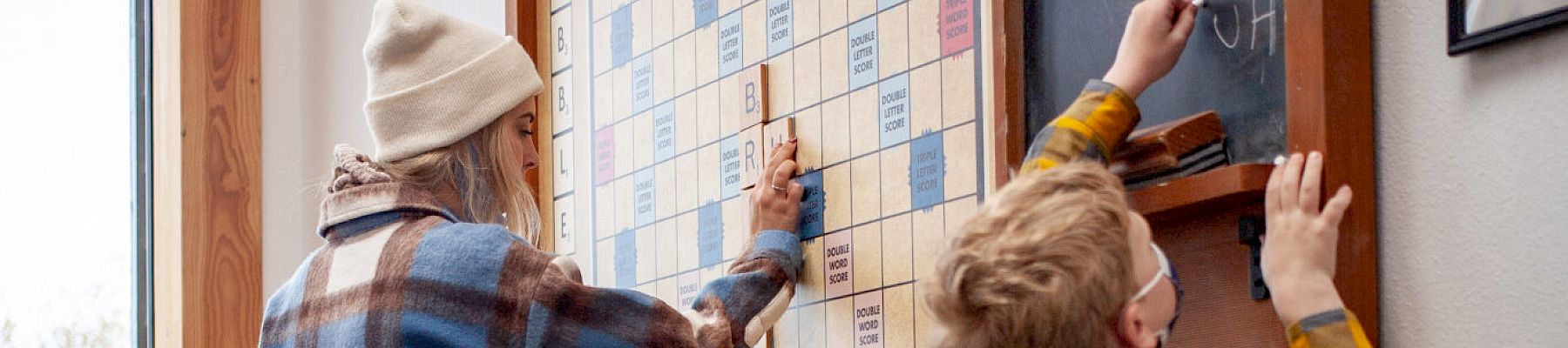 Three people, including two kids and an adult, play on a large wall-mounted Scrabble board.