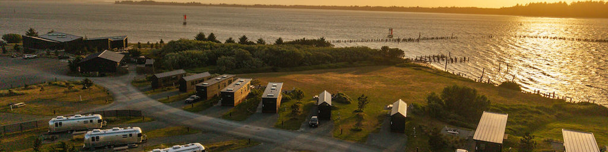 A scenic view of a lakeside RV park at sunset, with several trailers and cabins along a winding road near the water.