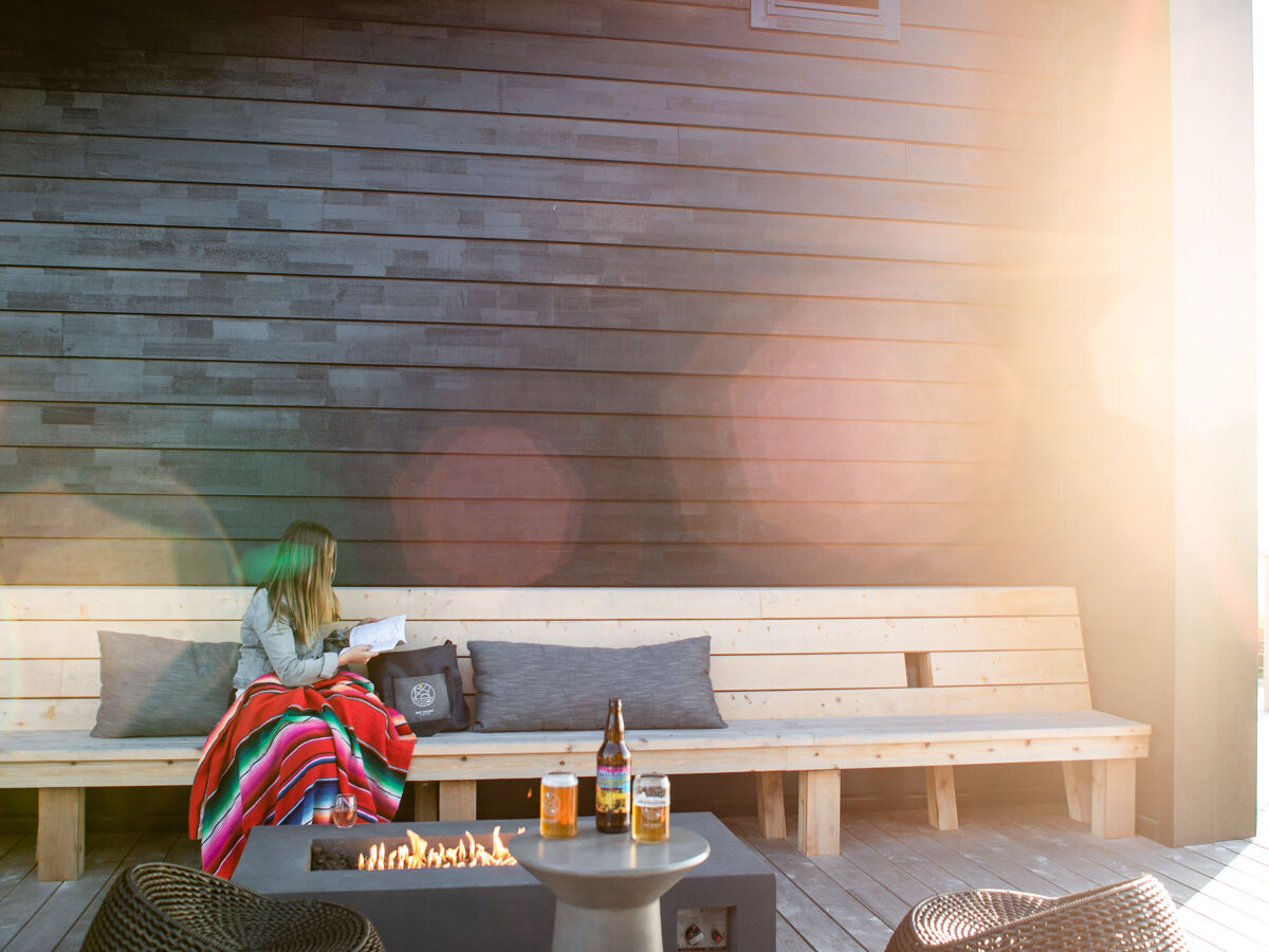 The image shows an outdoor patio with a person sitting on a bench beside a fire pit. Two round chairs and a side table are in the foreground.