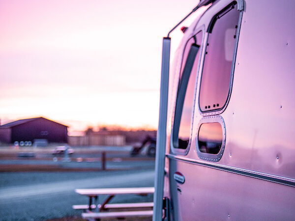 A silver RV is parked on a gravel path at sunset, with a picnic table nearby and a barn in the distance.