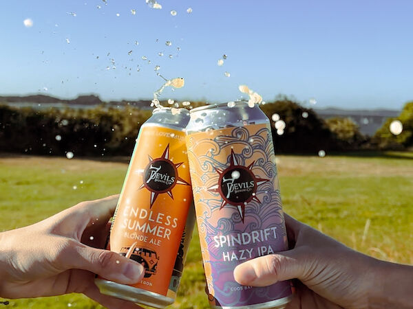 Two hands clinking cans of Spindrift beverages outdoors, with liquid splashing, in a grassy and scenic landscape.