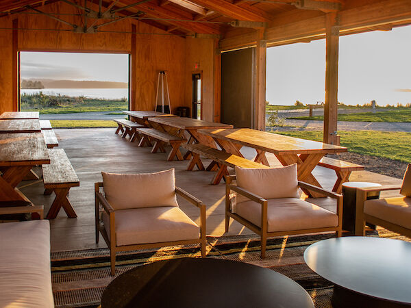 A cozy indoor space with wooden tables, chairs, and round black tables, leading to an open view of an outdoor area with grass.