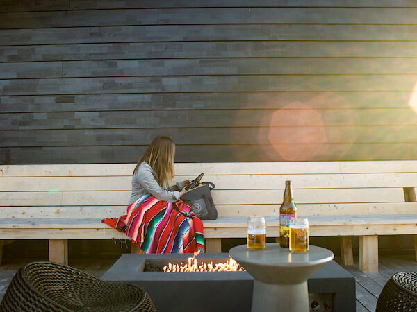 A person sits on a bench with a colorful blanket, near a table with drinks, and a fire pit outdoors.