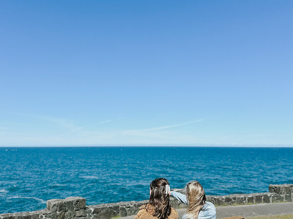 Two people sit on a bench facing the ocean under a clear blue sky.