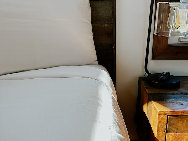 A neatly made bed with white sheets is beside a wooden nightstand featuring a vintage lamp and a small dish.
