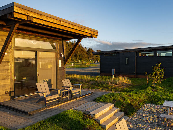 A small wooden cabin with a porch and two chairs at sunset. There's a gravel path and another cabin in the background under a clear sky.