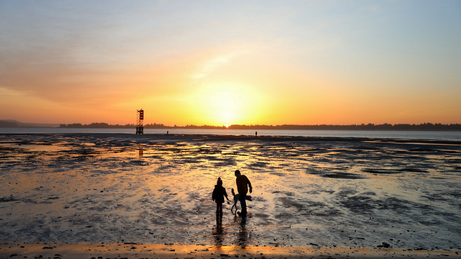 A sunset over a beach with two people walking on the wet sand, reflecting the colorful sky, and a lifeguard tower in the distance.