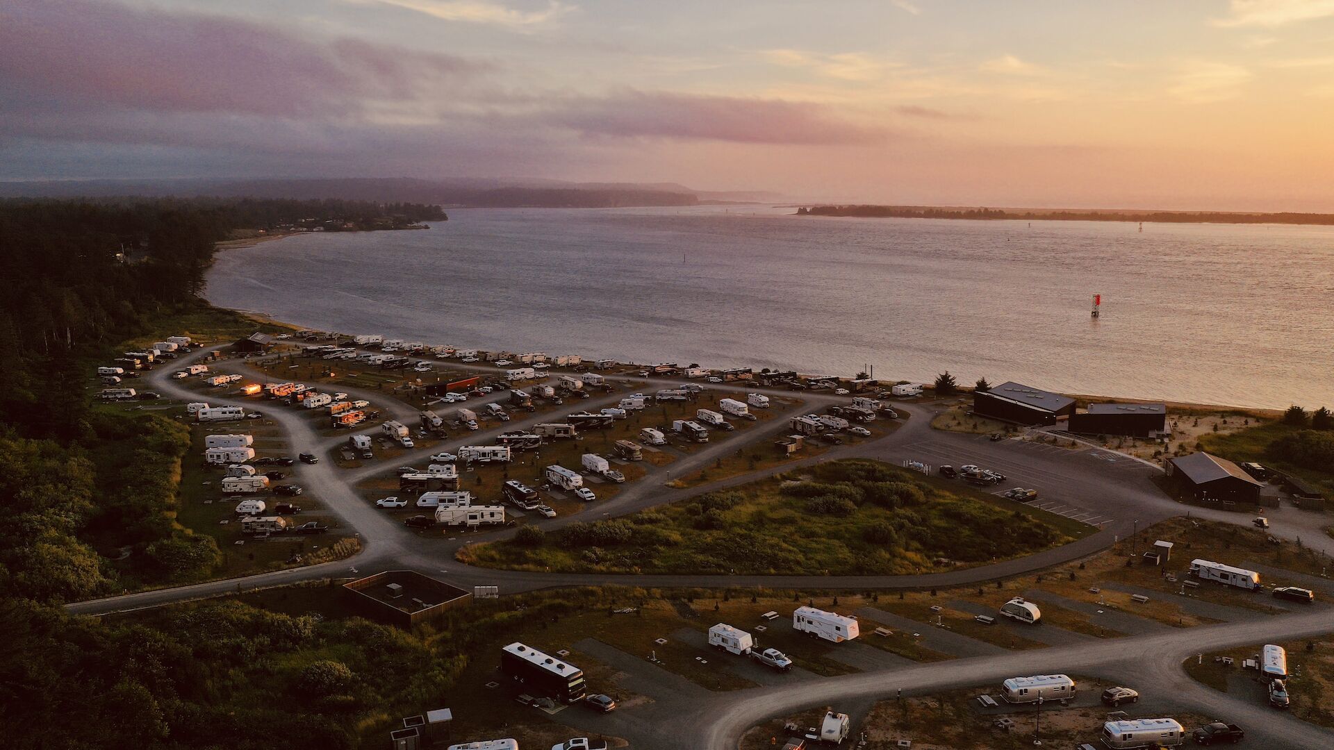 The image shows a coastal RV park with numerous campers and trailers near the shoreline during sunset, offering a peaceful and scenic view.
