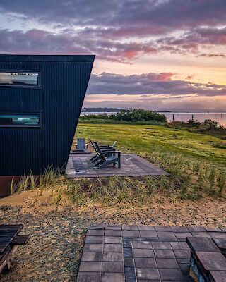 A modern building with a patio overlooks a grassy landscape and a sunset sky near the water.