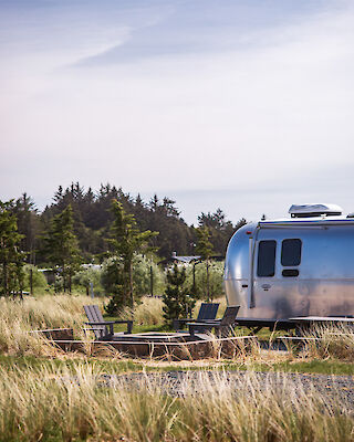 An Airstream trailer is parked in a grassy area with lawn chairs nearby and trees in the background under a clear sky.