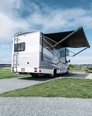 An RV is parked on a campsite near a grassy area and a body of water, with its awning extended under a blue sky with clouds.