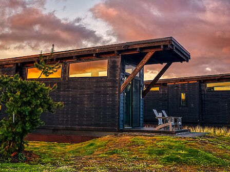 A modern cabin with a slanted roof at sunset, featuring large windows and outdoor seating, set against a dramatic sky.