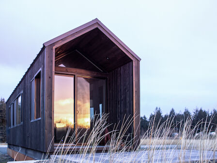 A small, modern cabin with large windows reflects a sunset, surrounded by tall grass and trees, under a cloudy sky.
