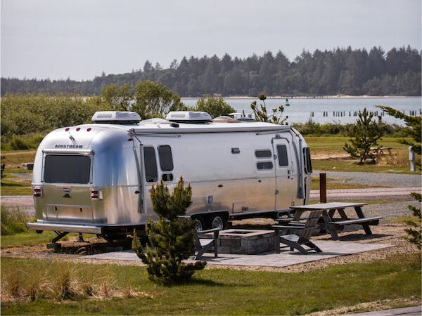 The image shows a silver Airstream trailer parked near a picnic table with a scenic view of a lake and trees in the background.