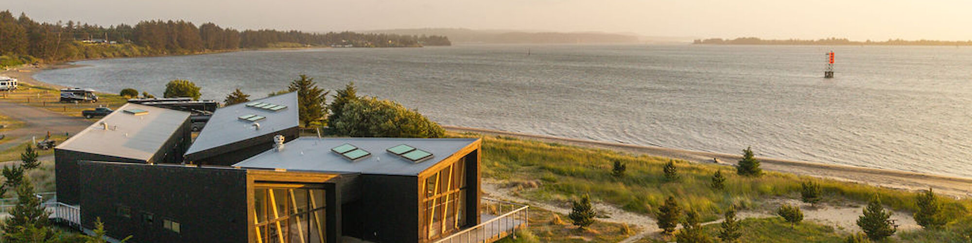 A modern building on the Southern Oregon Coast with greenery, pathways, and a calm body of water extending to the horizon at sunset.