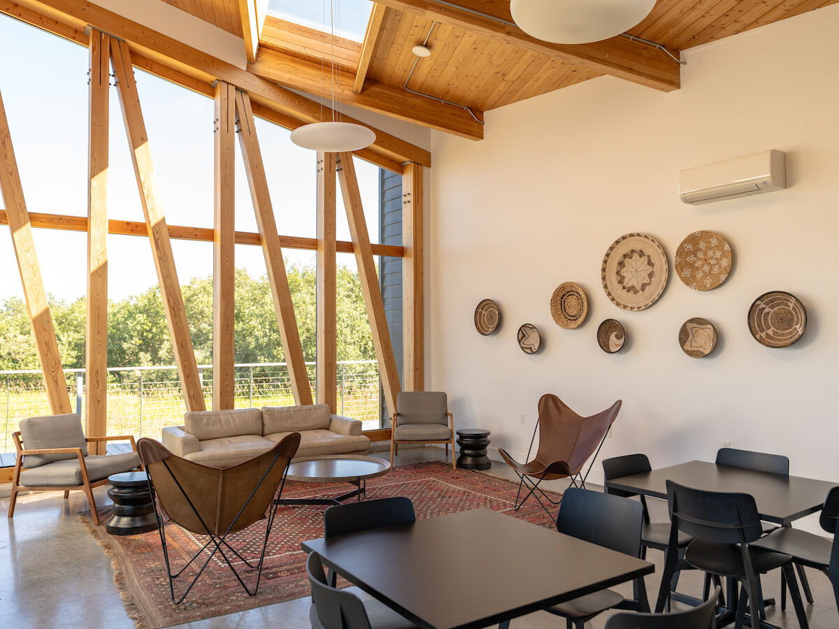 A modern room with large windows, wooden ceiling, wall decor, comfortable seating, and tables. Natural light enhances the cozy ambiance.