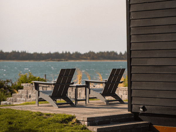 The image shows a serene scene with two chairs on a patio overlooking a body of water, next to a dark wooden structure.