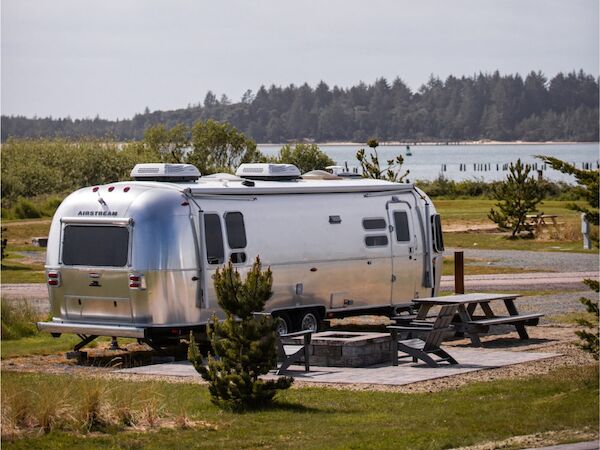 The image shows a silver Airstream trailer parked near a picnic table with a scenic view of a lake and trees in the background.