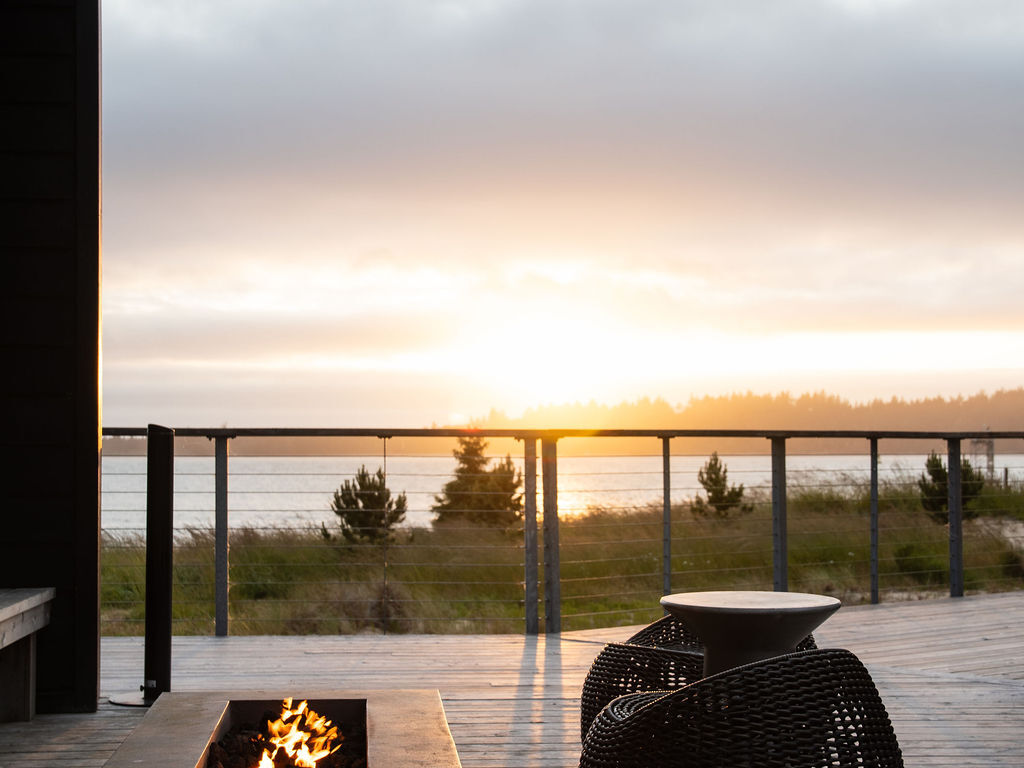 A modern outdoor patio at sunset with a fire pit, wicker chair, and glass railing overlooking a calm beach or lake, warm glow on the deck.