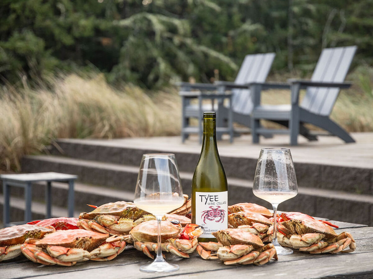 Crab feast setup with a bottle of white wine and two glasses on a wooden table, surrounded by sandy, coastal chairs and greenery.