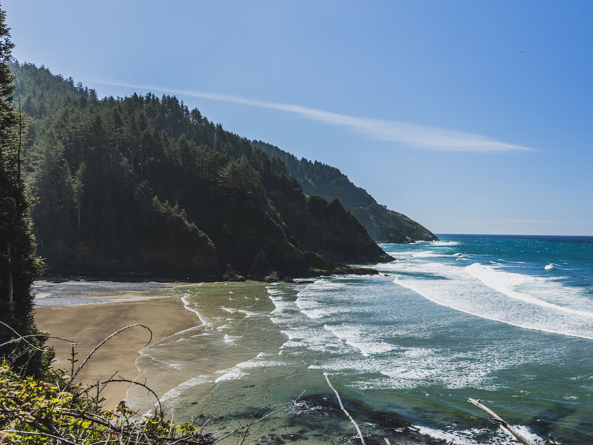 A rocky coastline with a sandy beach, forested cliffs, and blue ocean waves under a clear sky.