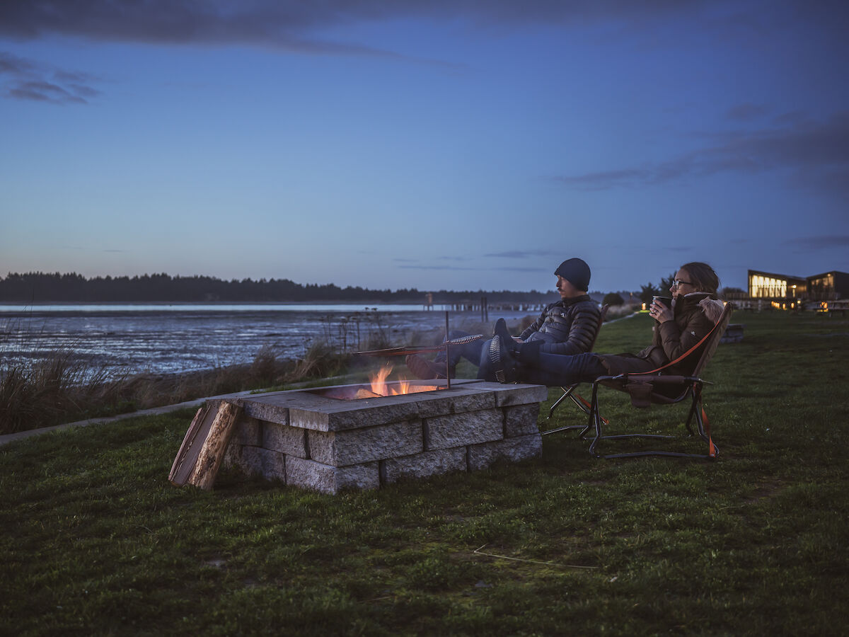 Two people sit by a small fire pit on a grassy shoreline at dusk, relaxing in chairs near the water.