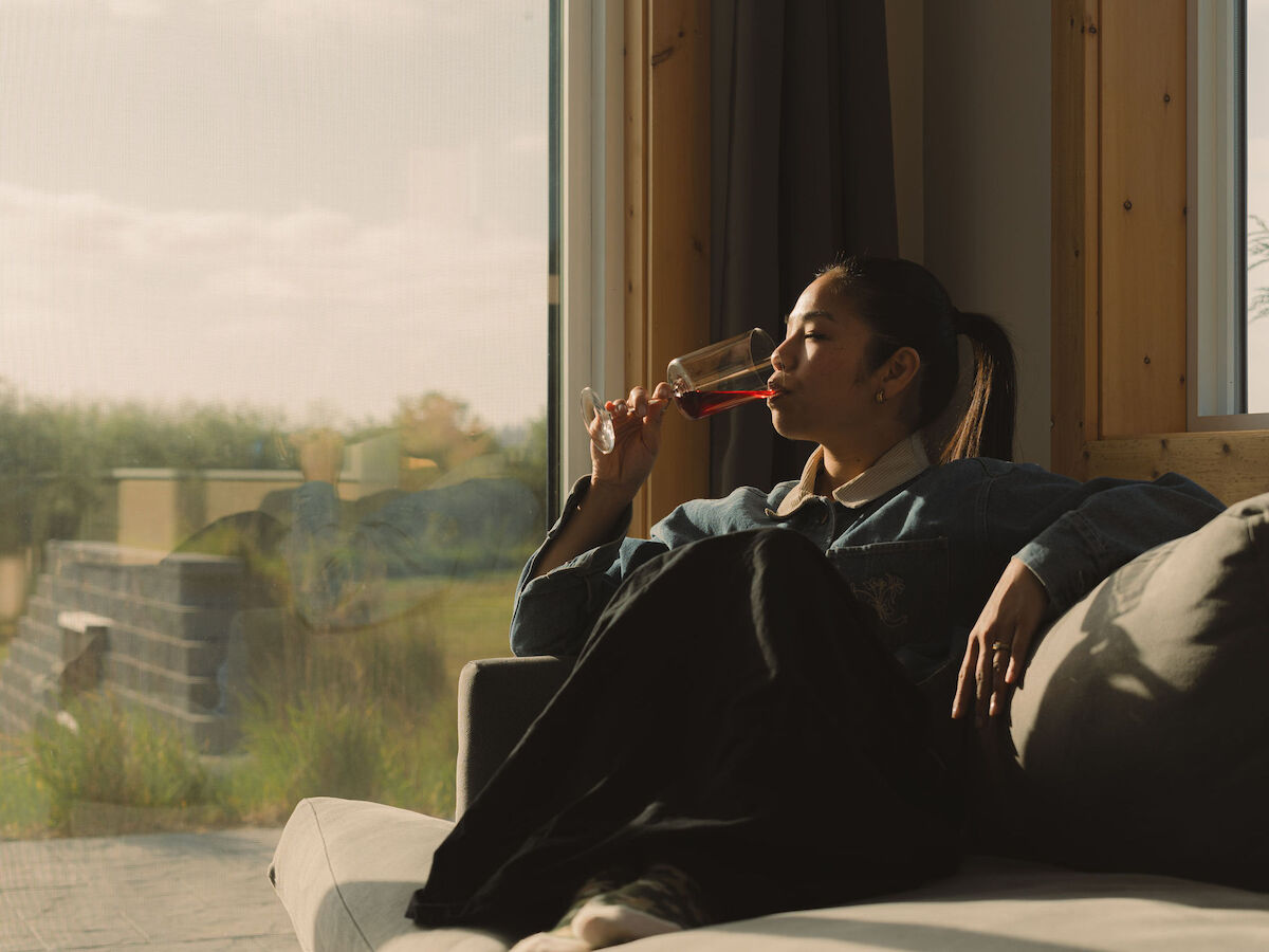 A person relaxes on a couch, gazing out a large window while drinking from a glass, with sunlight streaming into the room.