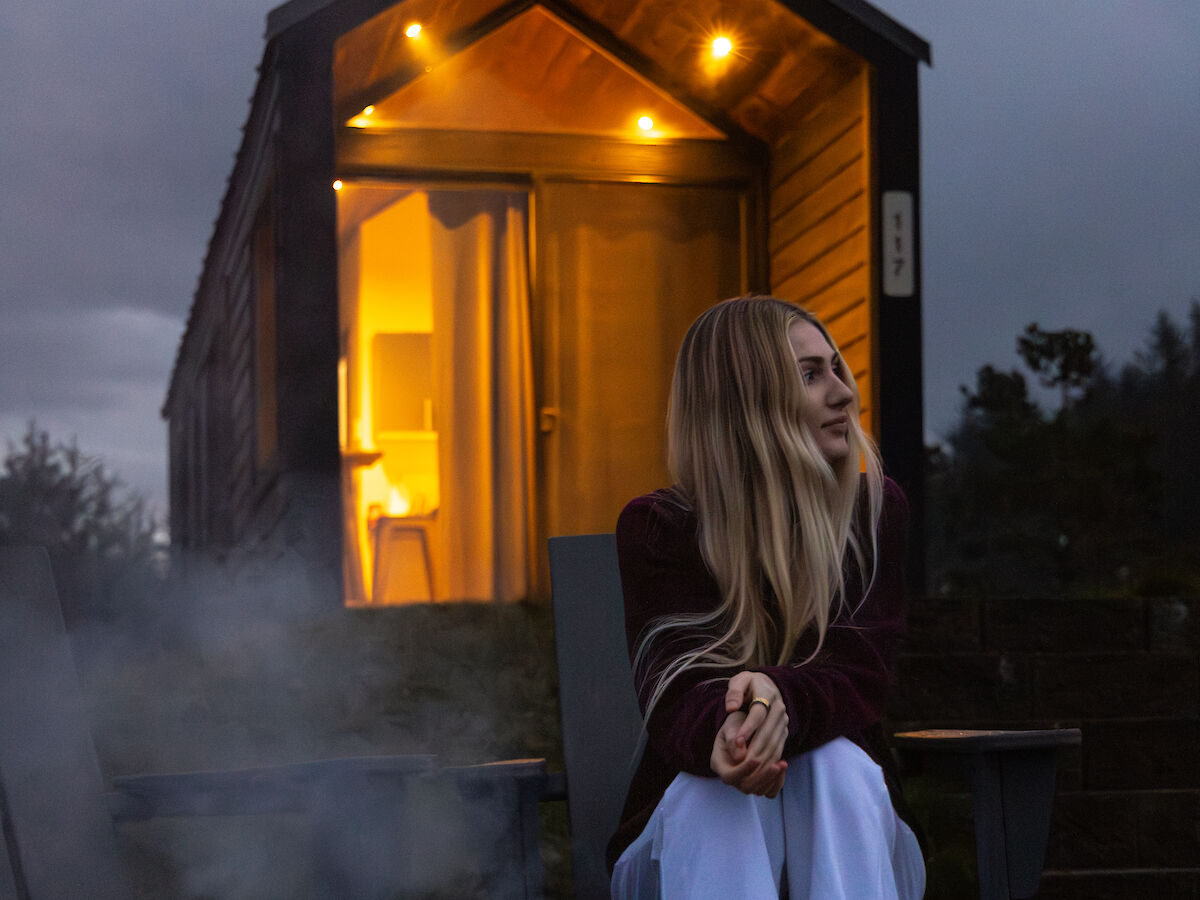 A woman sits by a warmly lit modern cabin at dusk, smoke rising from a nearby fire pit, creating a cozy, peaceful scene on the Oregon Coast.