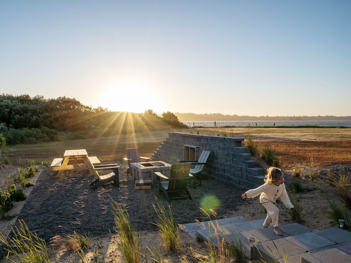 A child playing near a grassy seating area with picnic tables at sunset, surrounded by lush greenery and an open field.