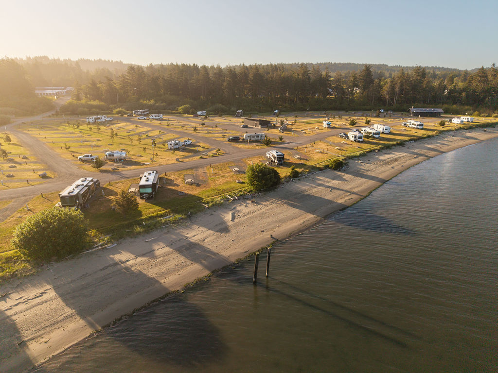 A lakeside campsite with RVs and tents spread across a grassy area, a sandy shoreline, trees in the background, calm water, and a sunny sky.