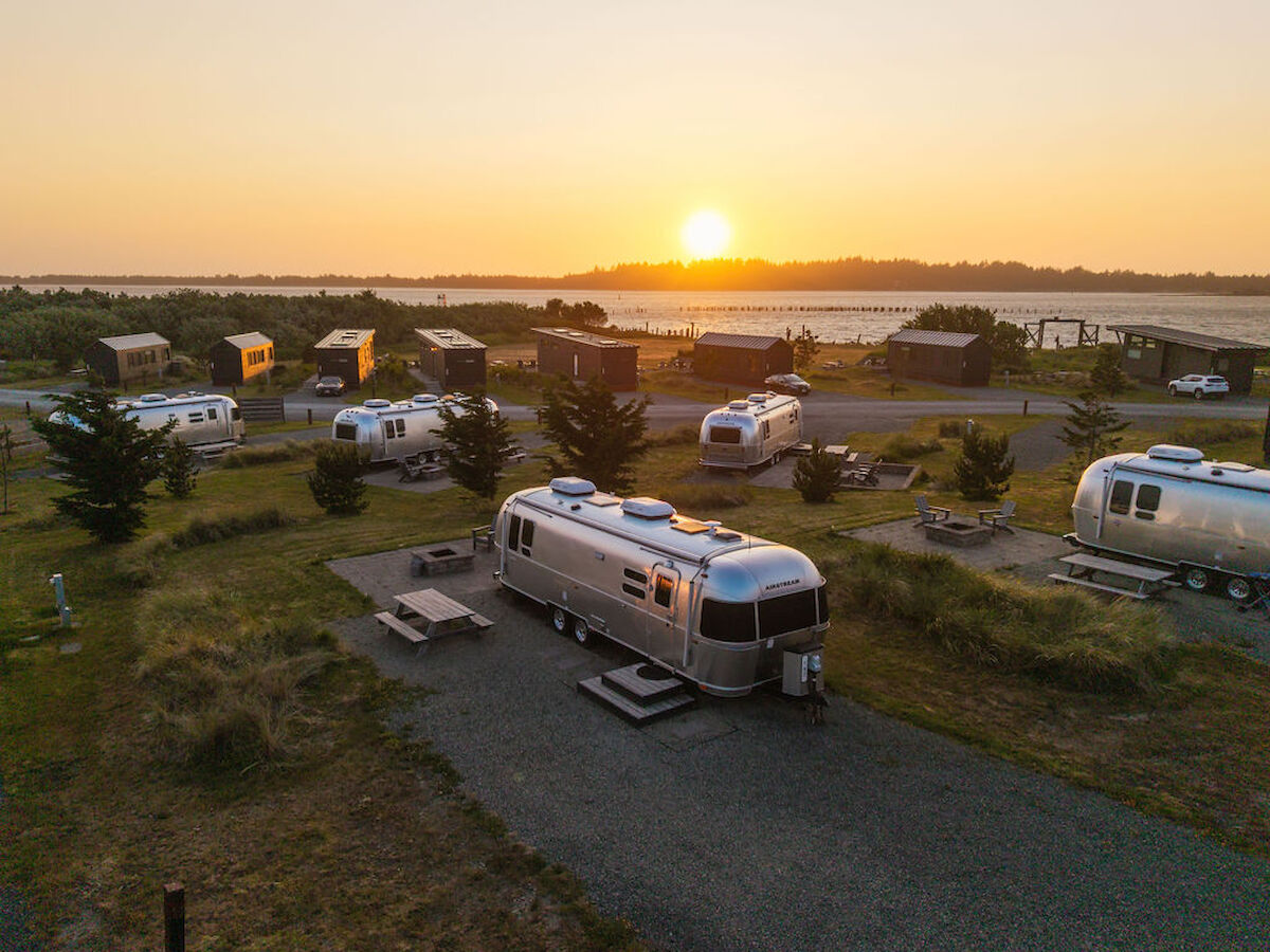 The image shows a sunset over a scenic RV park with several silver Airstream trailers, picnic tables, and surrounding greenery.