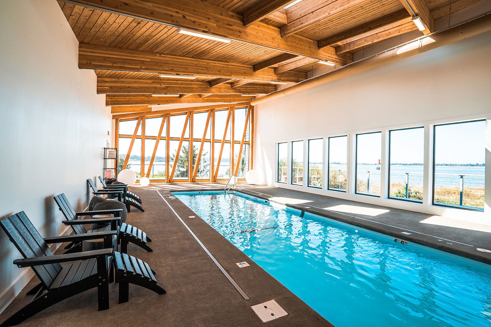 The image shows an indoor swimming pool with lounge chairs and large windows providing a scenic outdoor view.