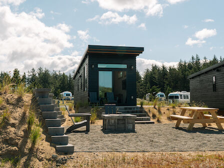 A small cabin with outdoor seating, picnic table, and nearby trailers under a cloudy sky surrounded by trees.