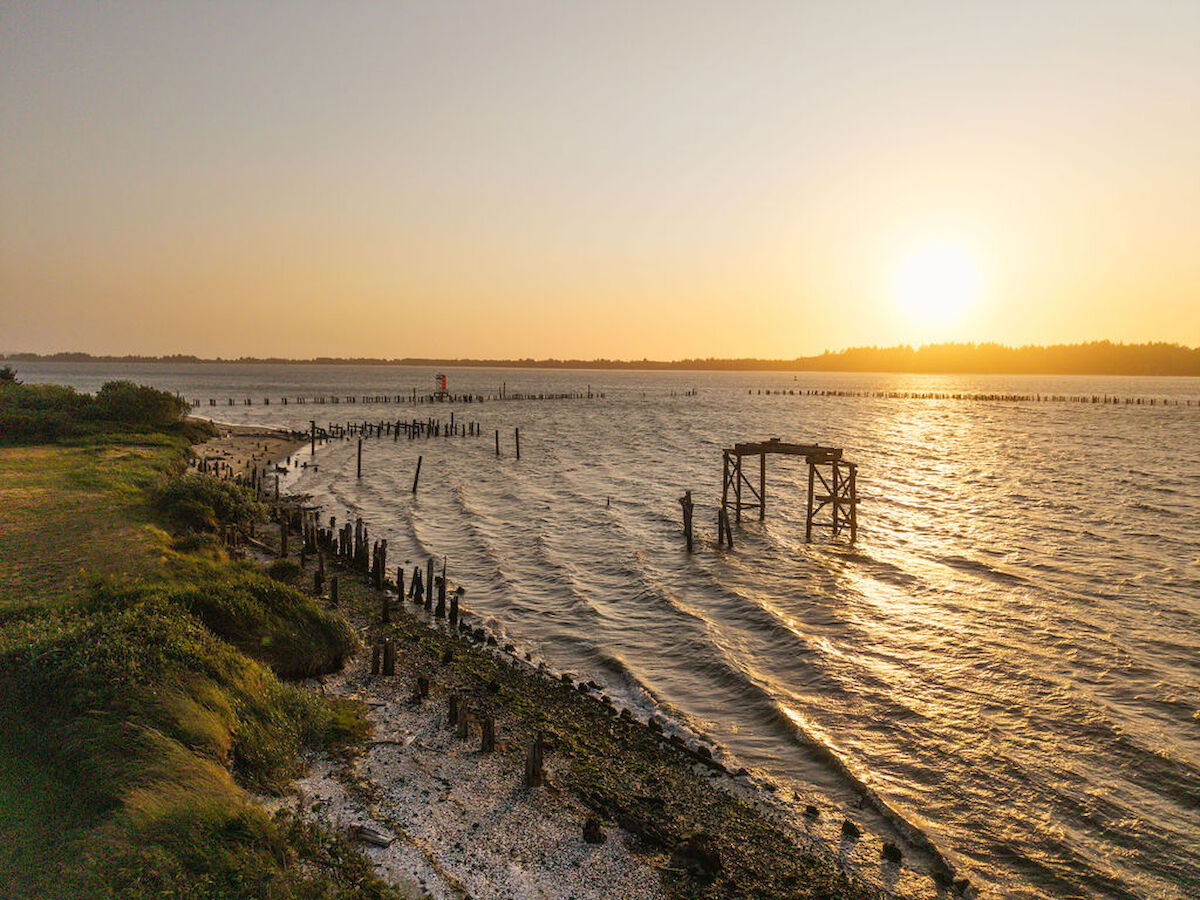 A serene sunset over a body of water with gentle waves, remnants of a pier, and grassy shoreline in the foreground.