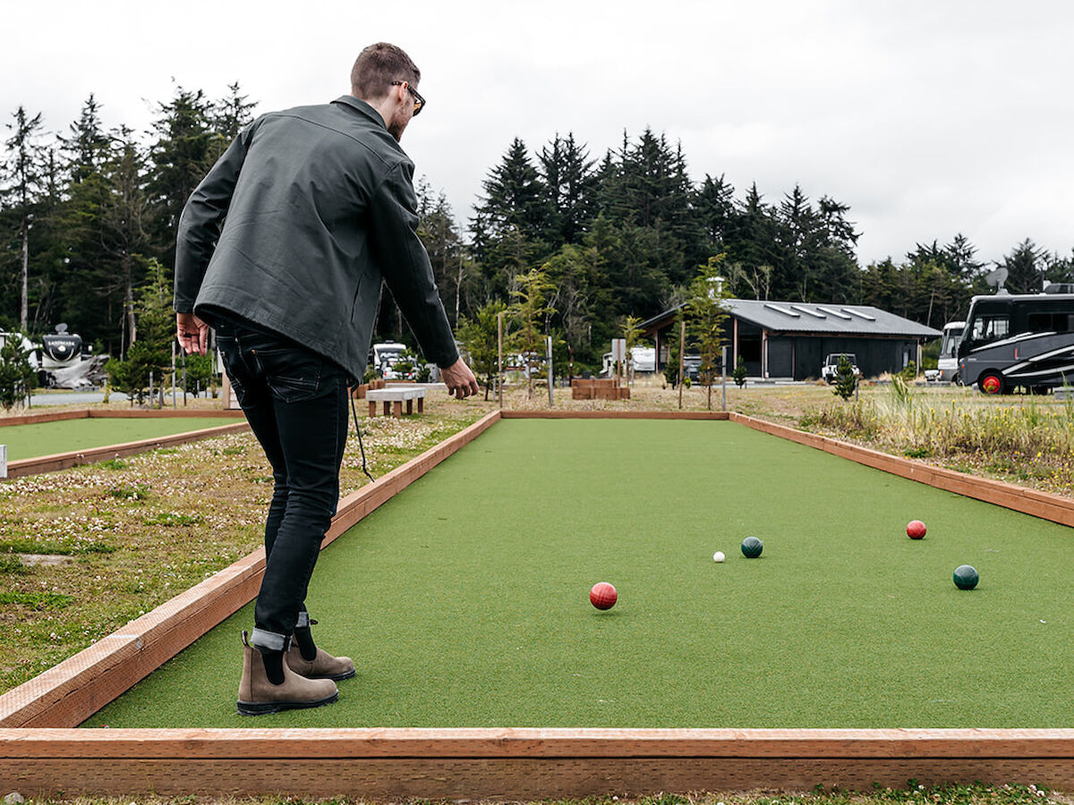 A person is playing bocce on a green outdoor court surrounded by trees and buildings under a cloudy sky.