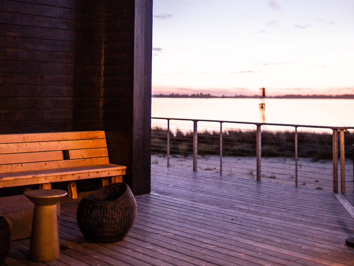 A wooden deck with a bench and small tables overlooks a serene body of water under a dusky sky.