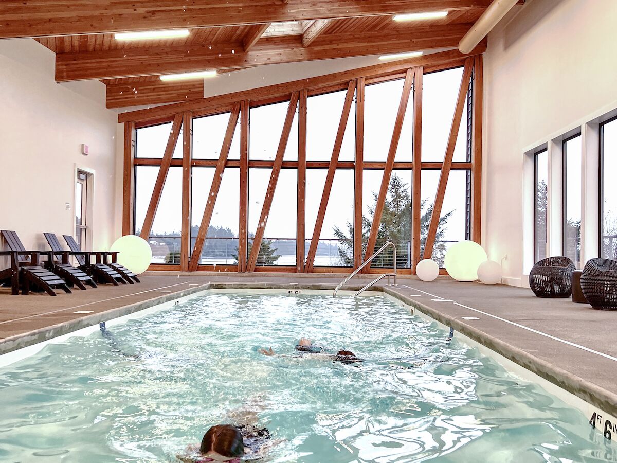 An indoor swimming pool with wooden beams overhead, large windows, and swimmers in the water, surrounded by lounge chairs on the deck.