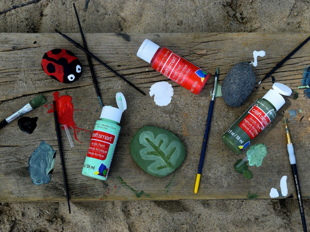 A creative craft scene with paint bottles, brushes, a ladybug toy, a green leaf stamp, clay pieces, and colorful art supplies on a wooden surface.