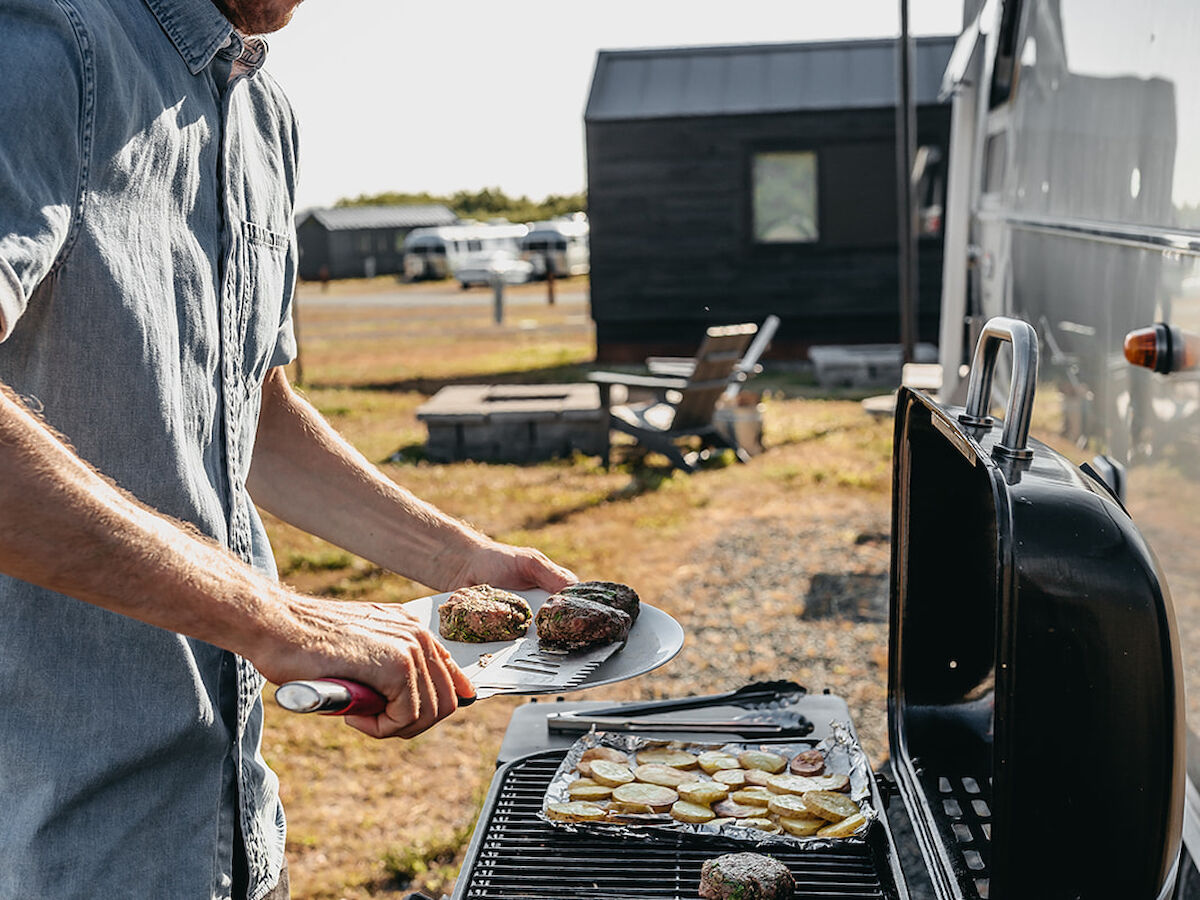 A person is barbecuing outdoors next to a modern trailer, with cooked food on the grill in a casual setting.