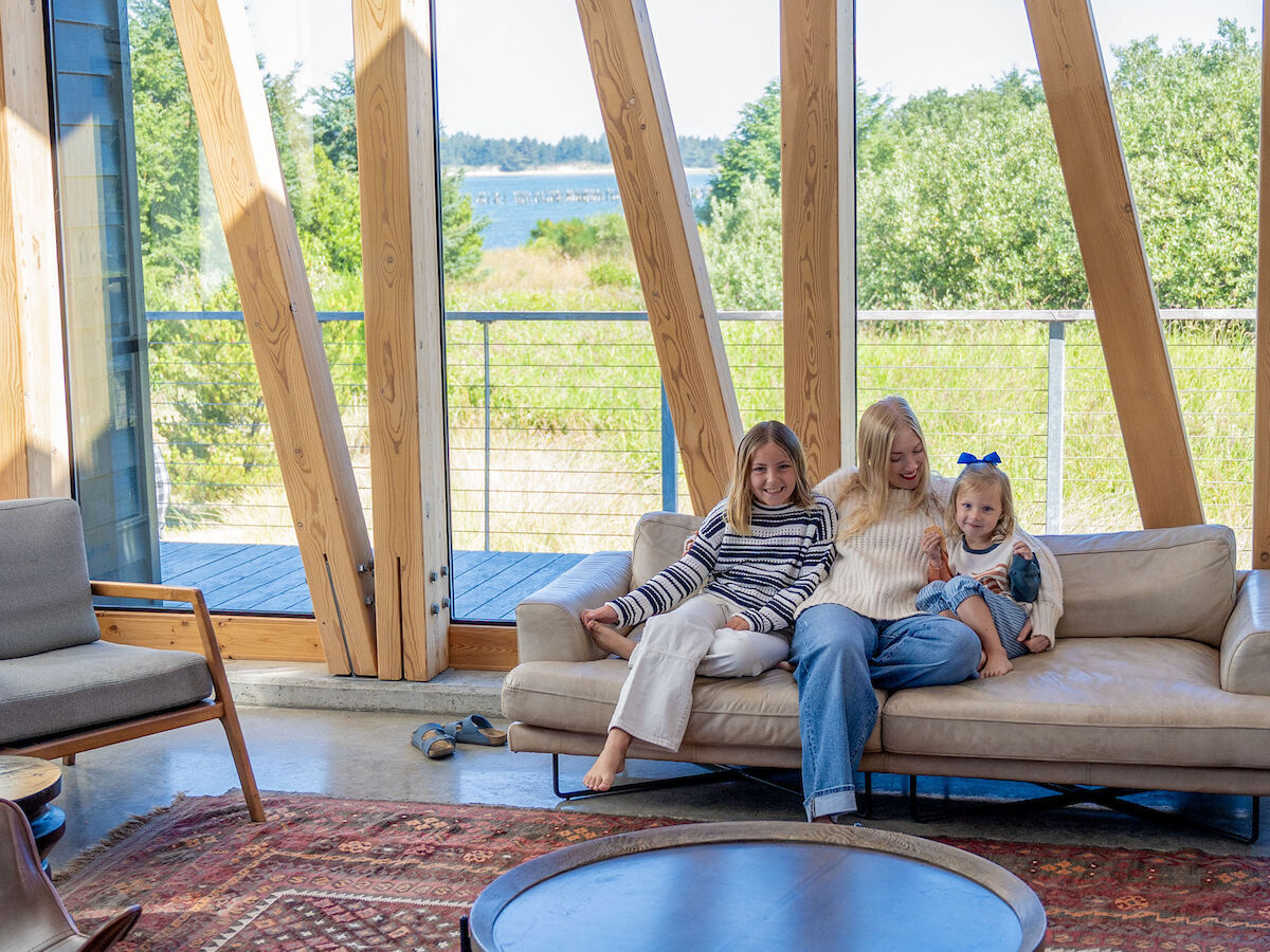 A cozy living room with large windows, a sofa, a round table, and a scenic view outside. Three people sit on the sofa.