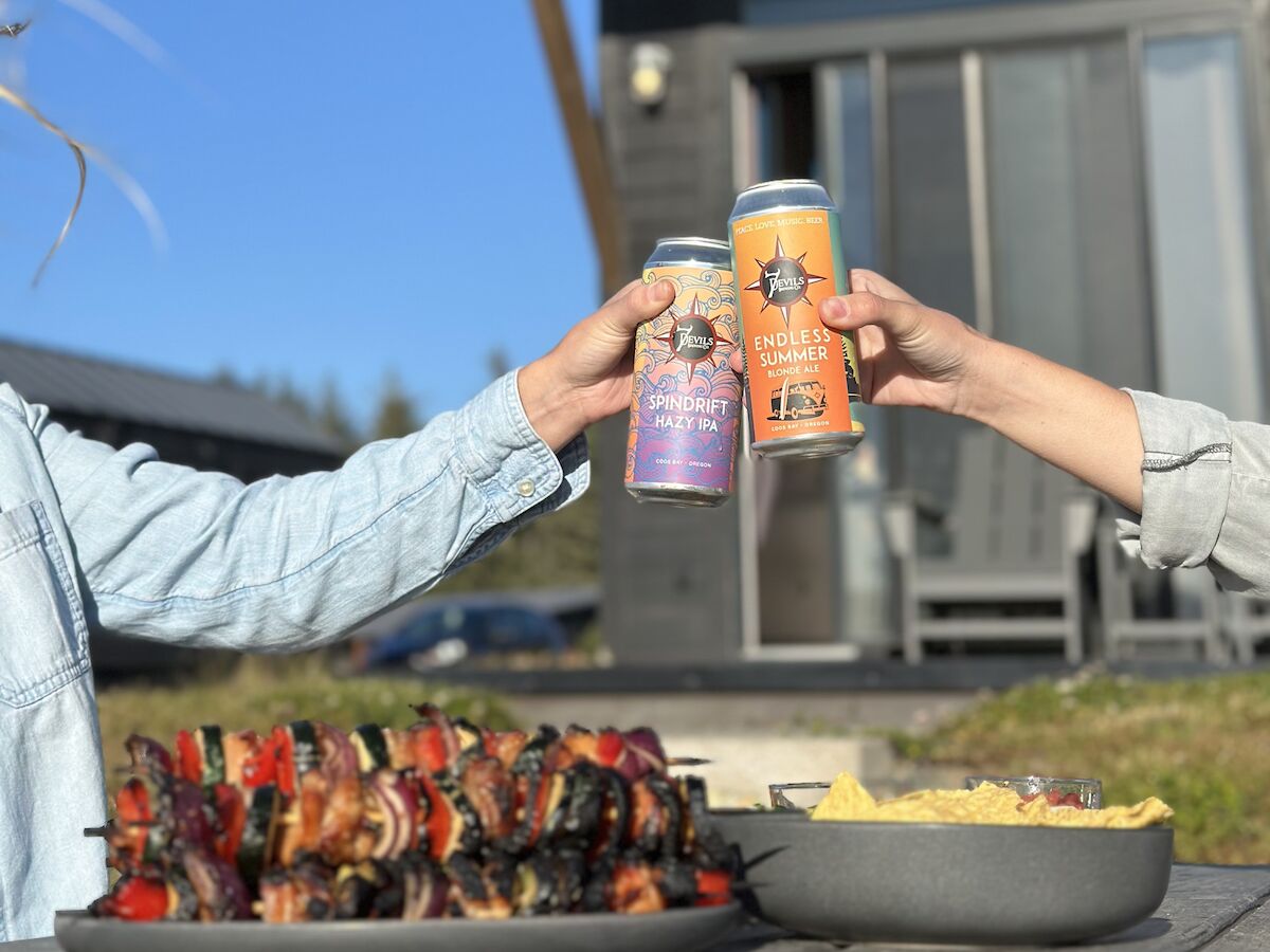 Two people toast with cans of beer near a cabin. Plates of grilled skewers and chips are on the table in the foreground.