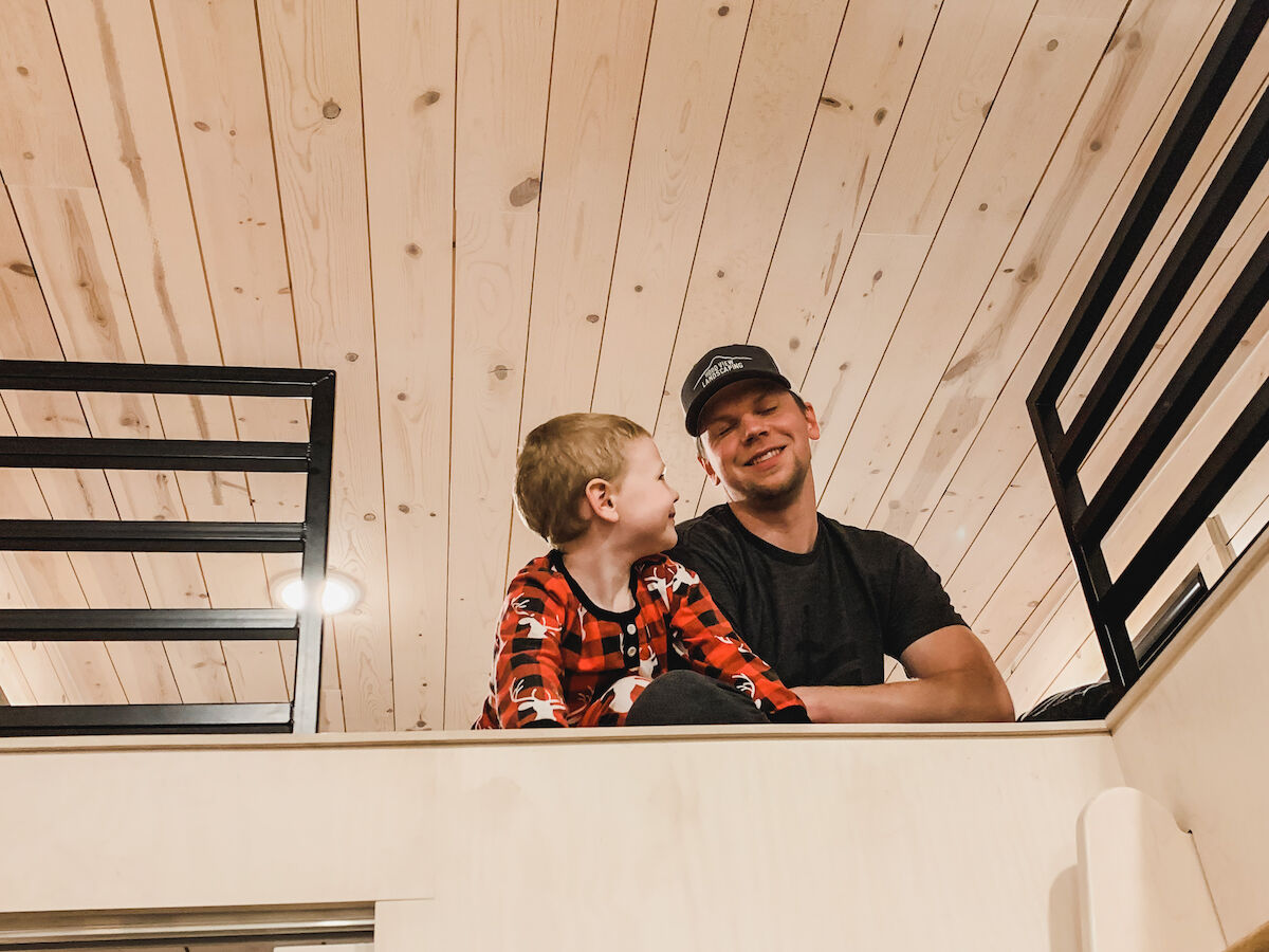 A child and an adult sit on a loft area with wooden paneling, looking at each other and smiling.