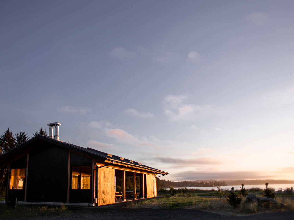 A wooden house is set against a scenic backdrop of trees and a river under a broad sky at sunset.
