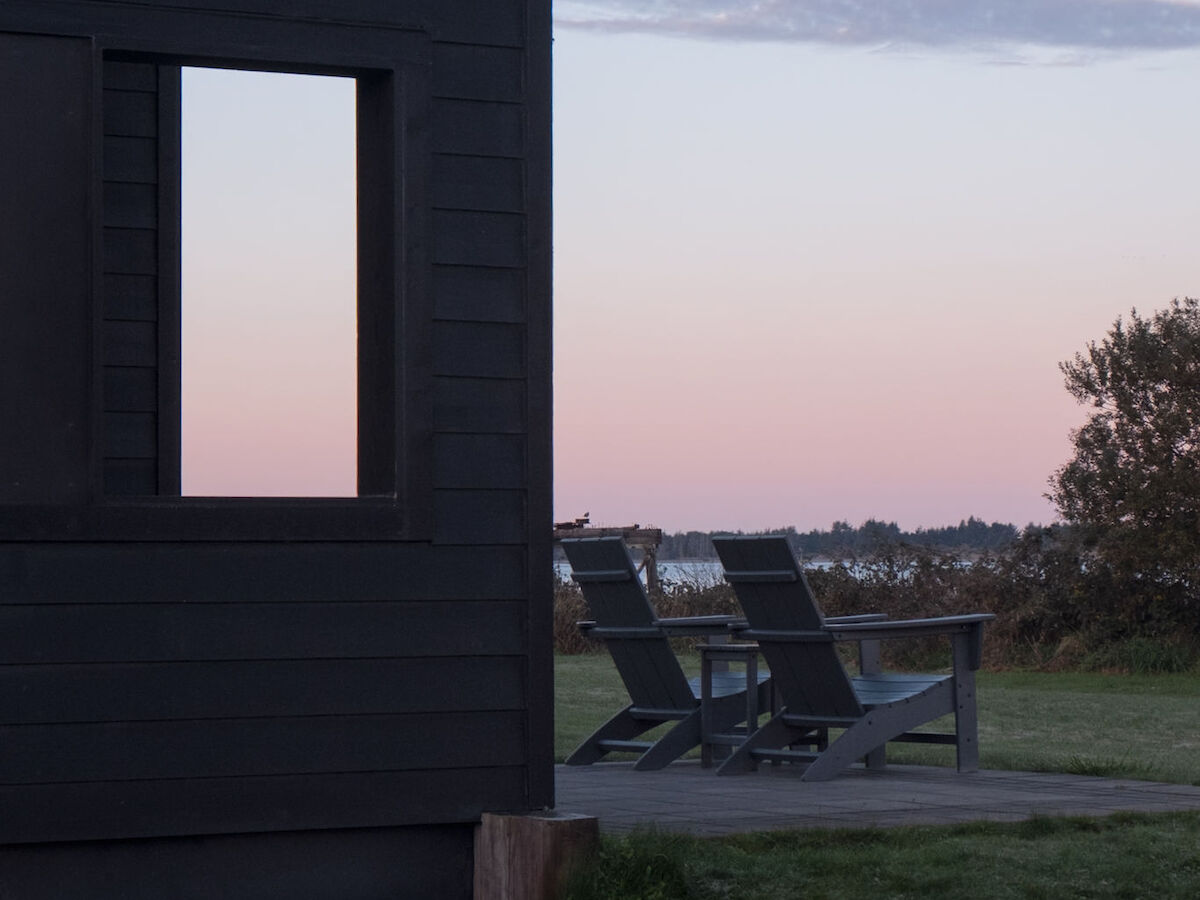 A black wooden building in a grassy area with chairs on a deck, set against a serene sunset sky.