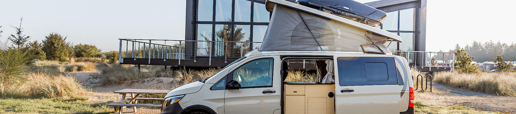 A camper van with an open pop-up roof is parked near a modern building with large windows, in a grassy area.